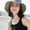 Une femme souriante porte un bob chapeau gris avec ficelle sur un décor clair