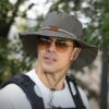 chapeau-aventurier-homme-style Un homme style explorateur porte un chapeau aventurier avec des grosses lunettes dans un décor de nature