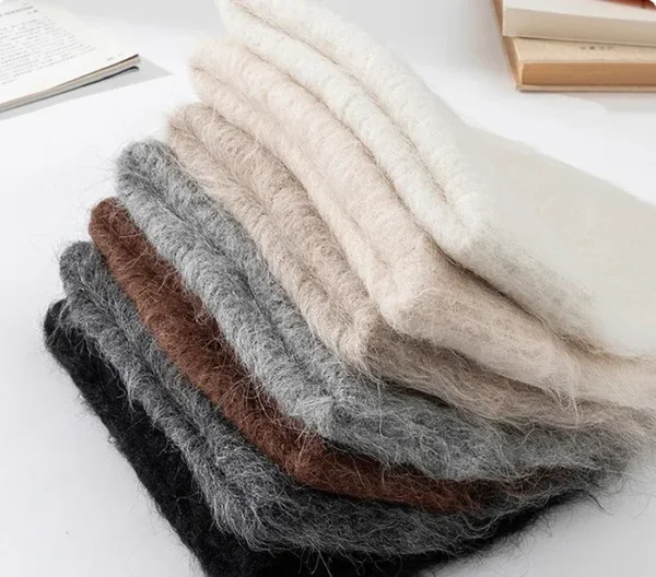 bonnet-laine-naturelle-empiles-couleurs-variees Pile de bonnets en alpaga pliés, montrant différentes teintes naturelles comme beige, gris, marron et noir, soulignant la douceur de la laine d’alpaga.